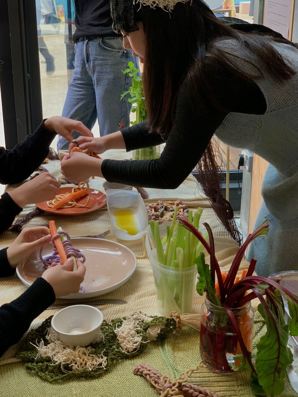 A woman leaning across a table of vegetables, edible yarn and knitted pieces while guests tie knots