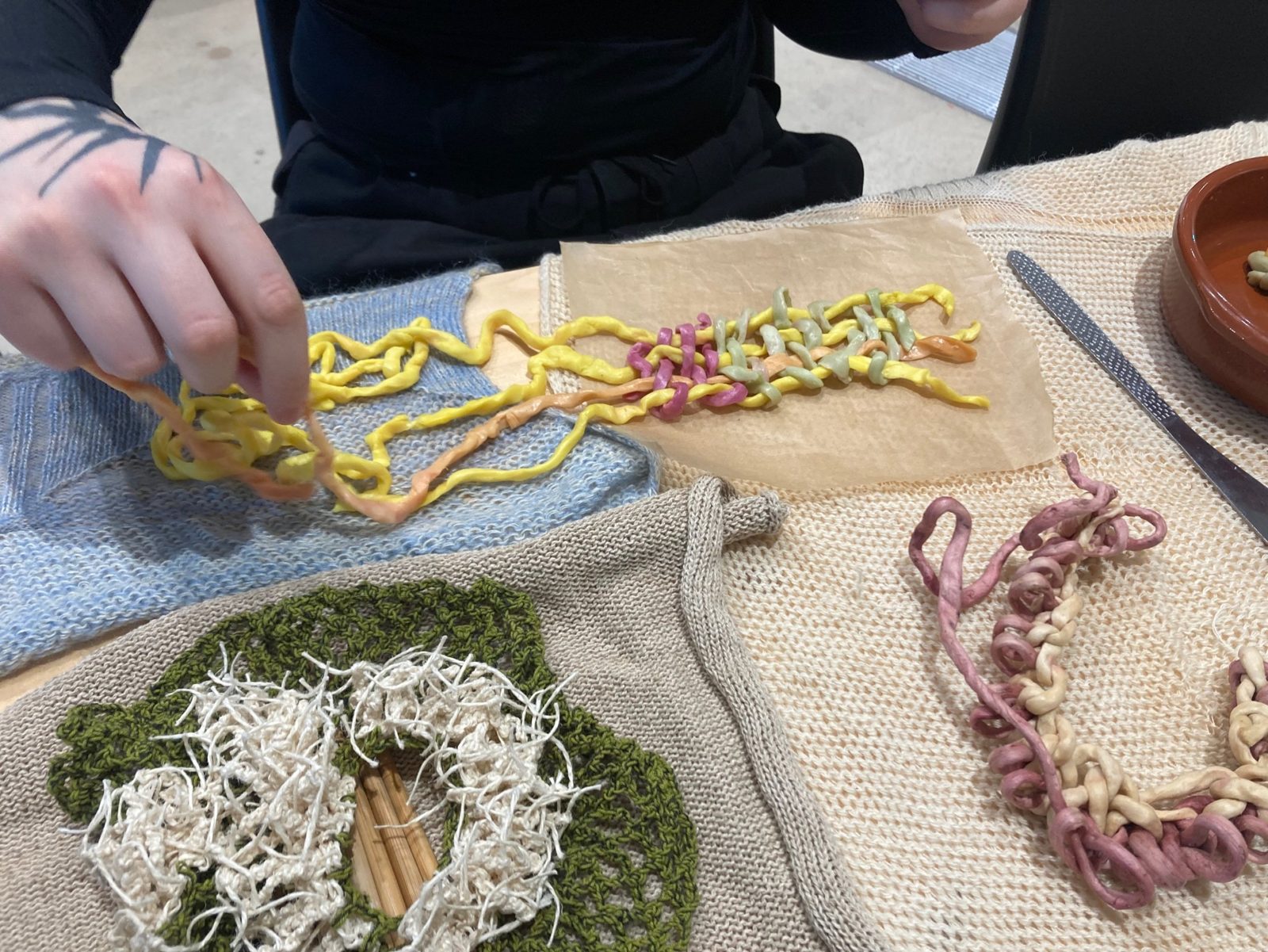 Hands knotting yellow and orange edible yarn on a blue knit, beside a woven yarn sample and green crochet