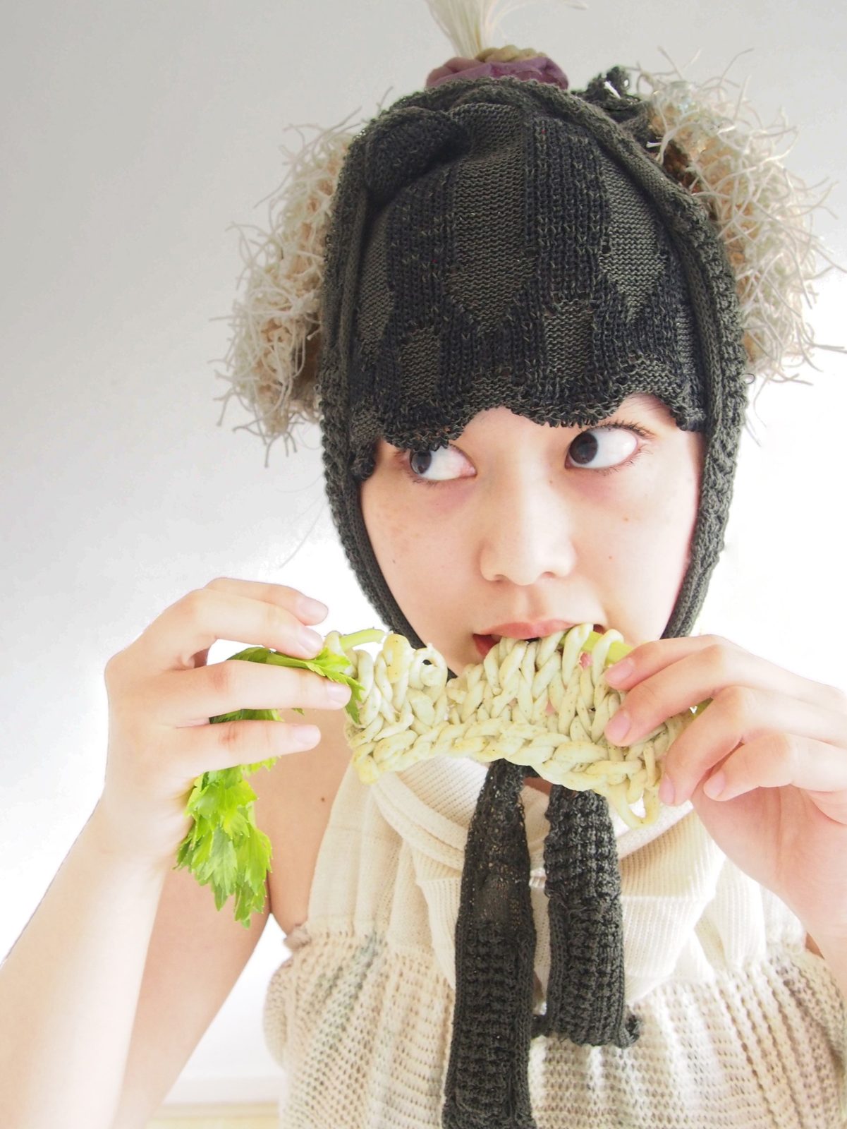 A woman in a knitted hood biting a knitted celery loop
