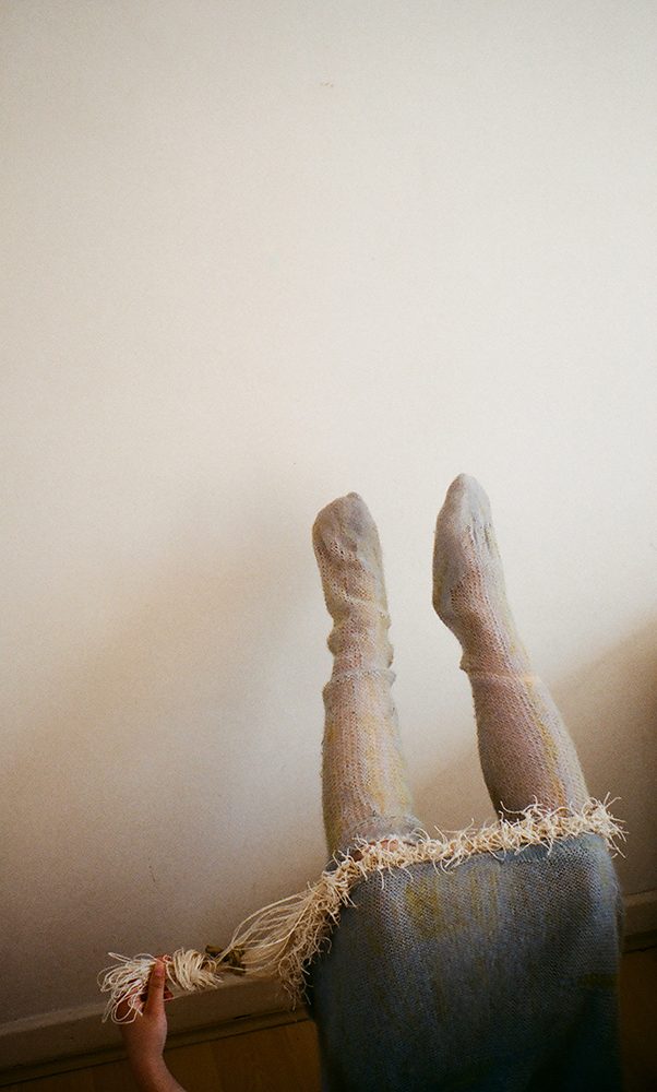 Legs in knitted socks raised against a wall beside the fringed hem of a knitted dress
