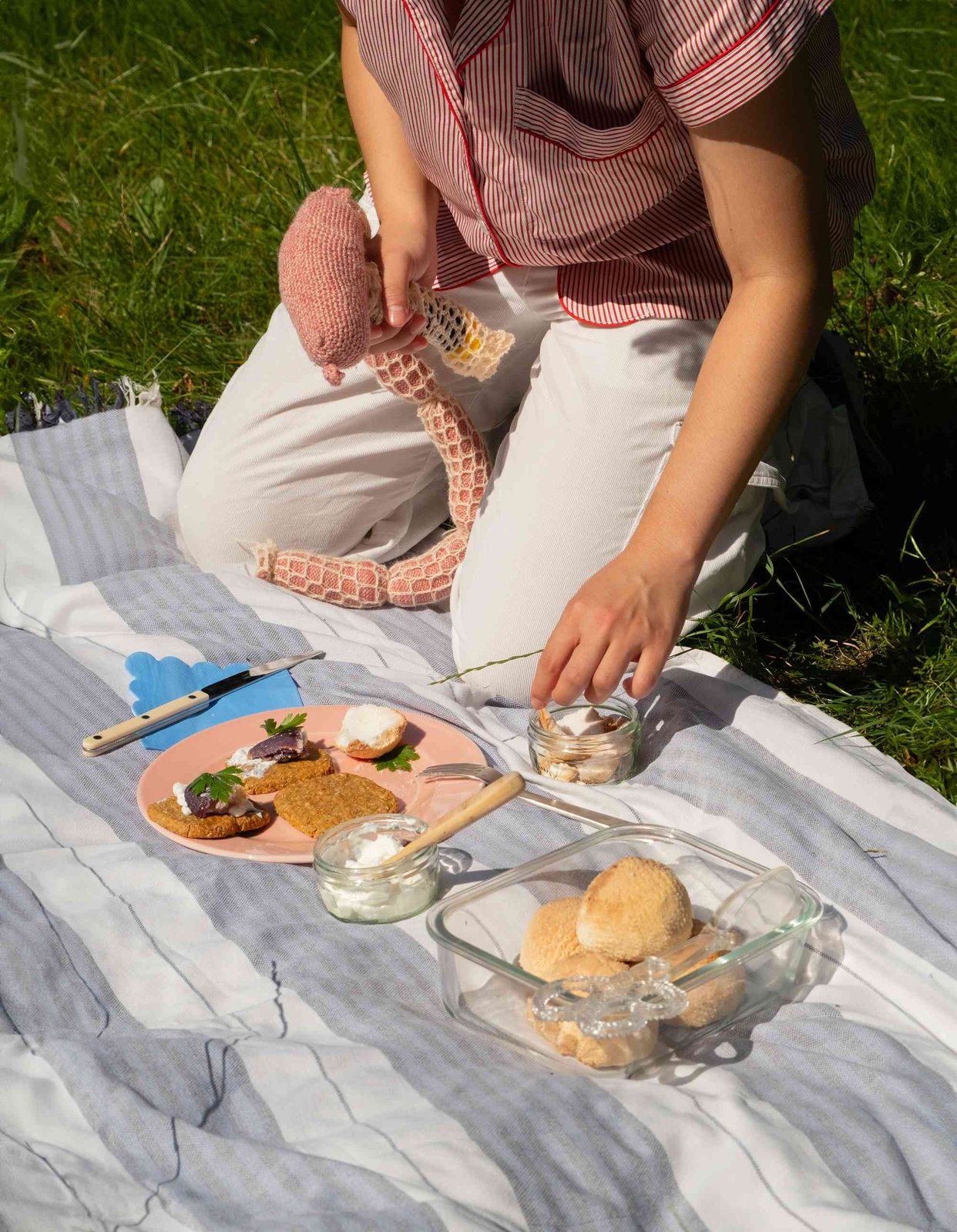 The performer kneeling on a picnic blanket with a knitted sausage hammer, a plate of food and a glass dish of crocheted mochi balls