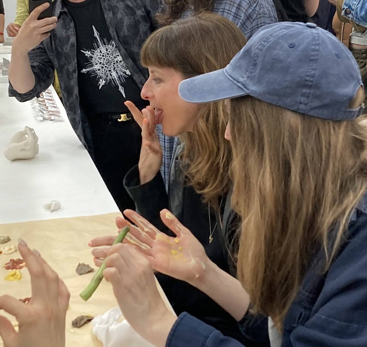 A visitor tasting paint from her hand at the long table, with another visitor in a blue cap