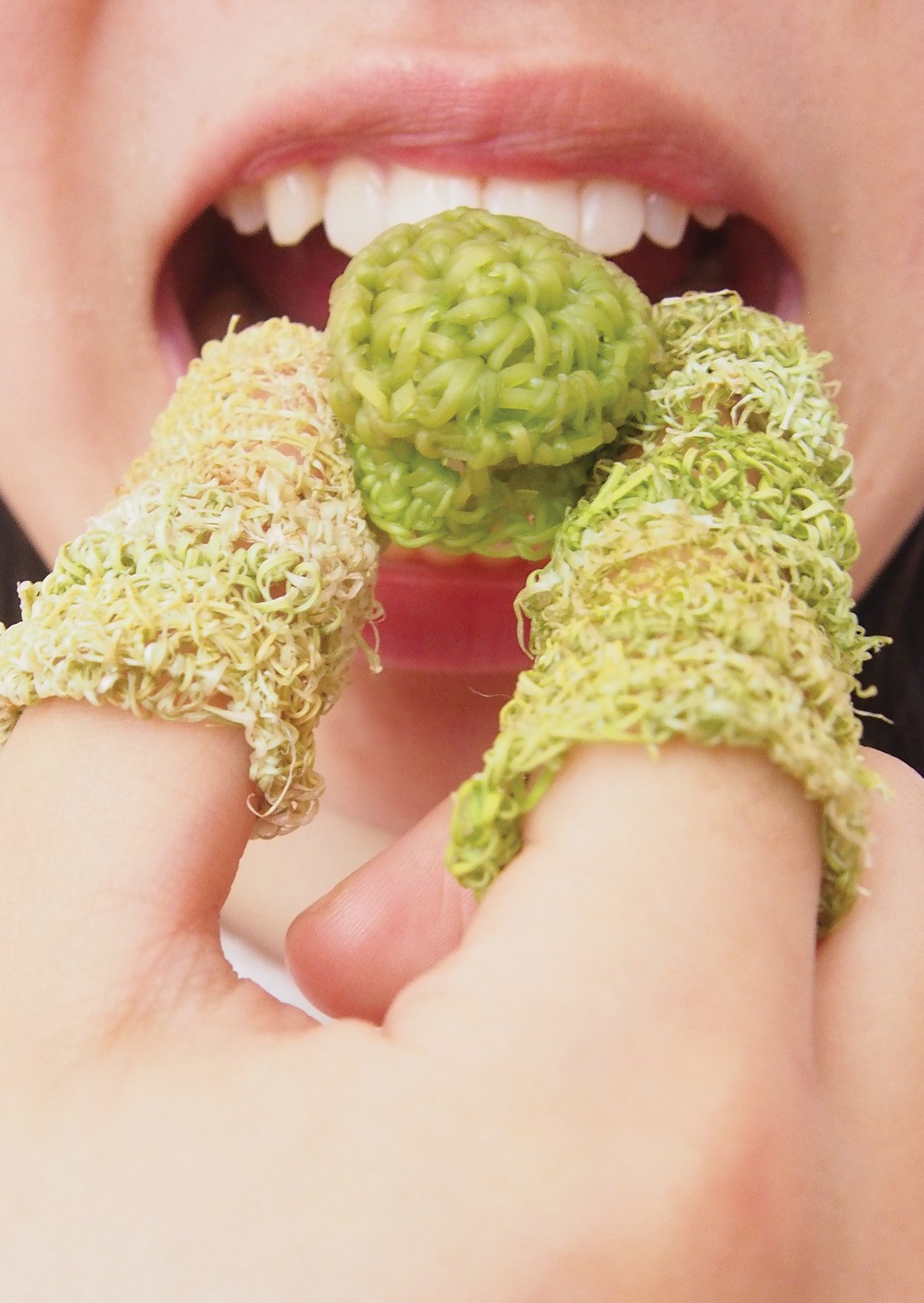 A mouth about to bite a knitted green edible ball, with knitted vegetable yarn rings on two fingers