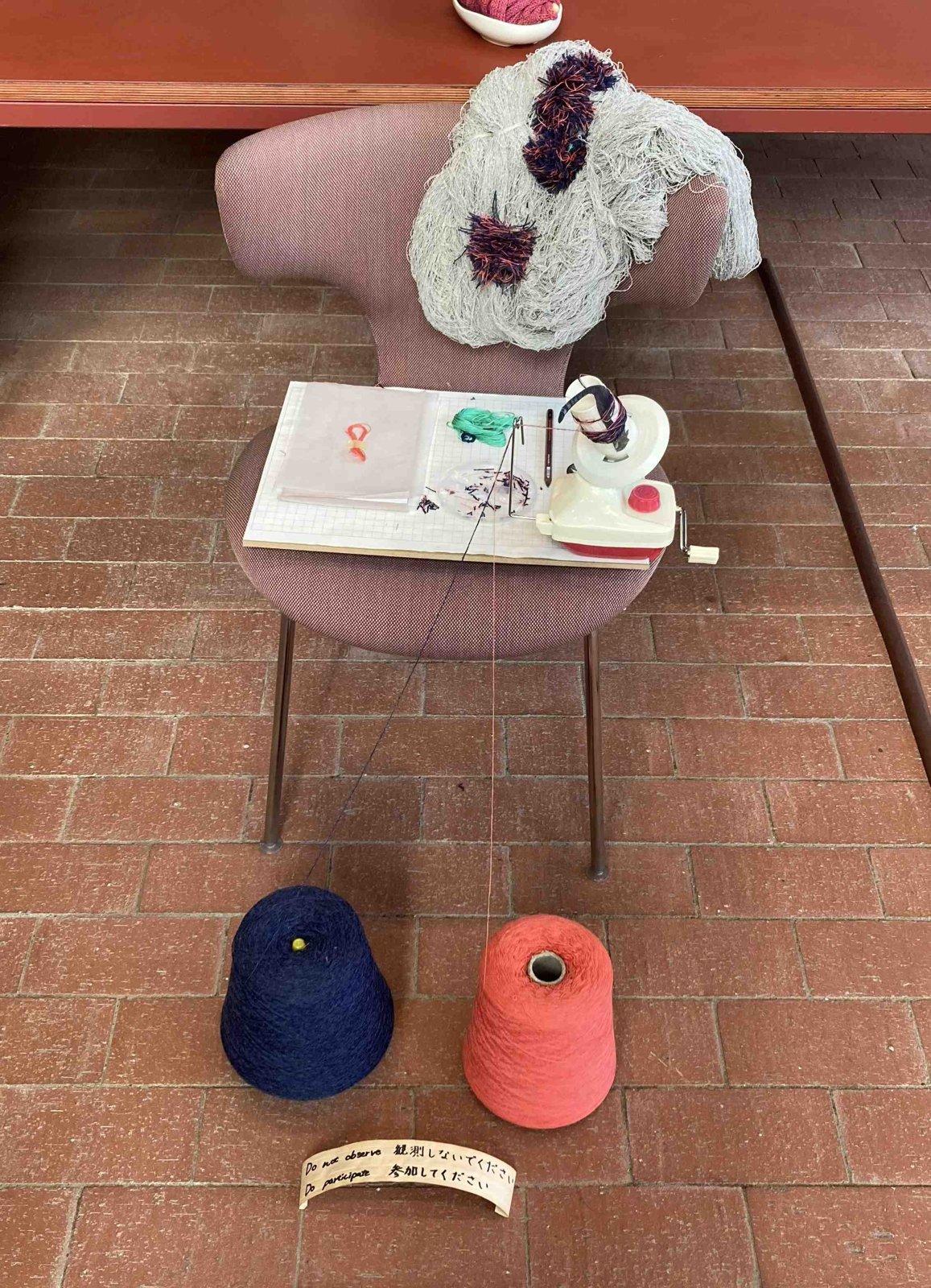 A knitted textile draped on a chair beside a hand yarn winder, blue and red cones of yarn on a brick floor