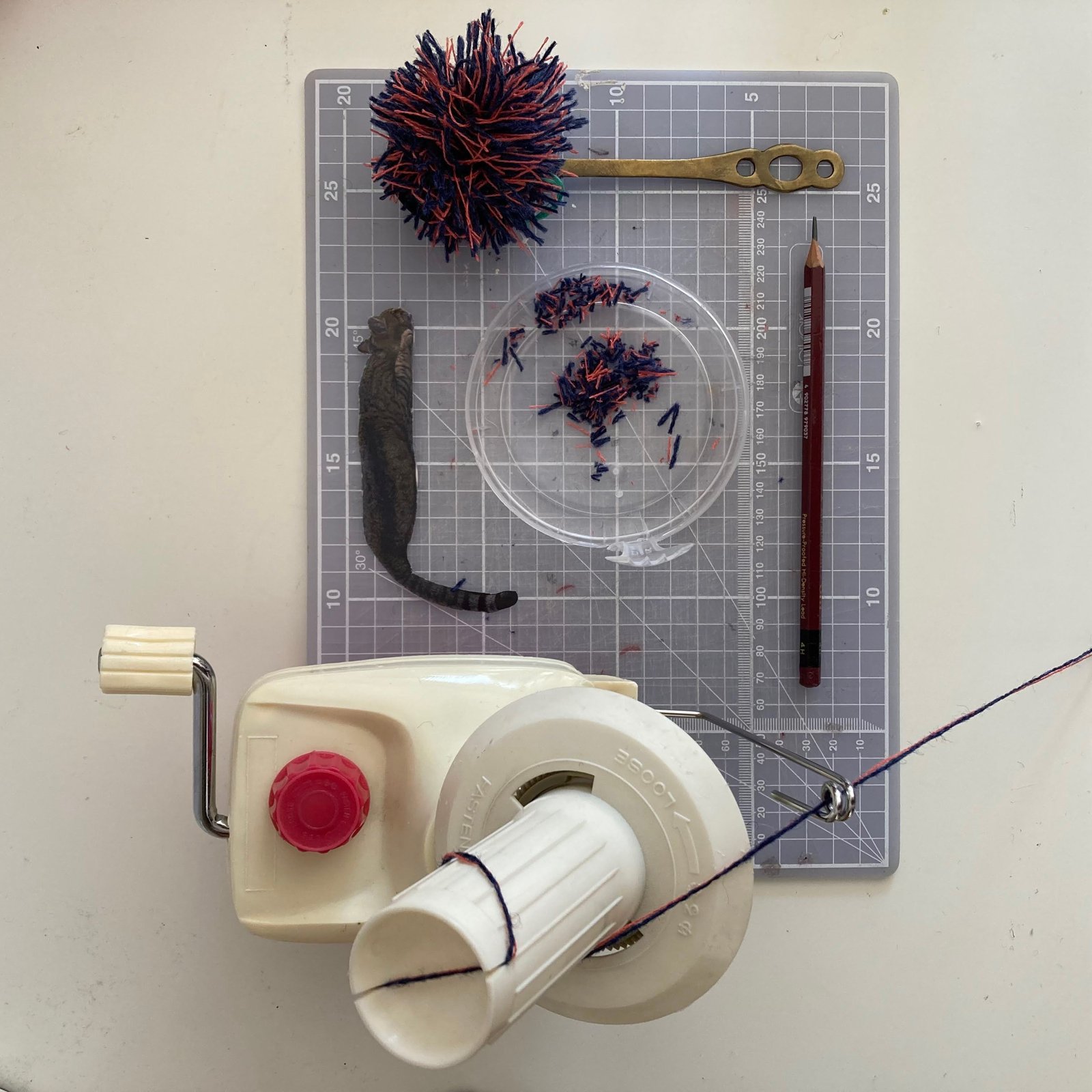 Hand yarn winder on a cutting mat with a pom-pom, purple threads in a glass dish and a cat-shaped textile